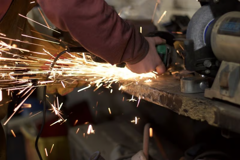 A person uses an angle grinder to cut metal, creating a bright spray of sparks in an industrial Rust Belt setting.