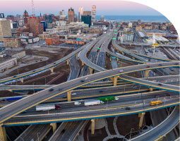 Multi-level highway interchange with traffic at dusk against a city skyline.