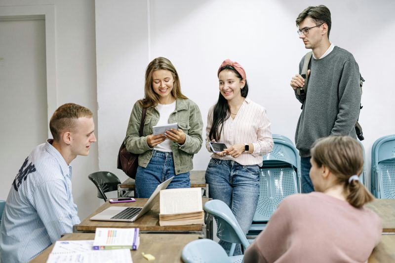 Five students collaborate at a desk with a laptop and open book during a 2024 college study session.