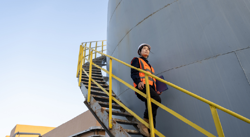 15 Worker in hard hat and safety vest climbing a metal spiral staircase beside a large industrial storage tank.
