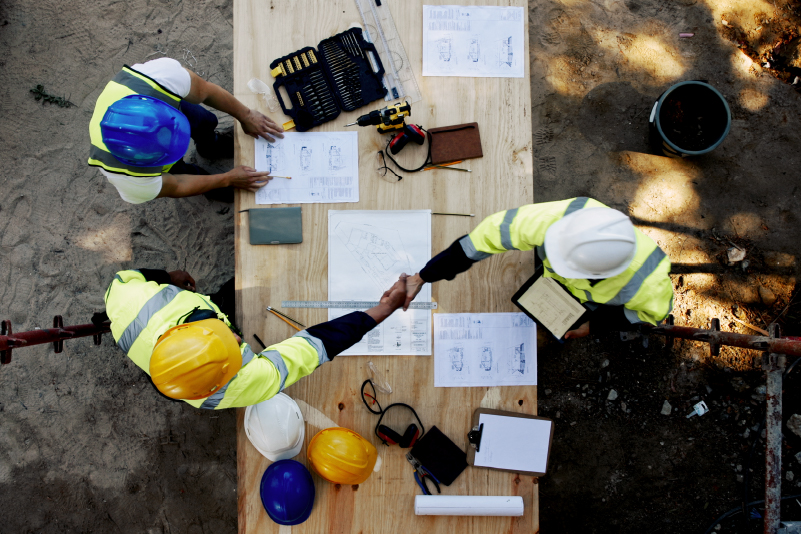 5 Construction workers in high-visibility vests shake hands over a wooden table with blueprints and tools.